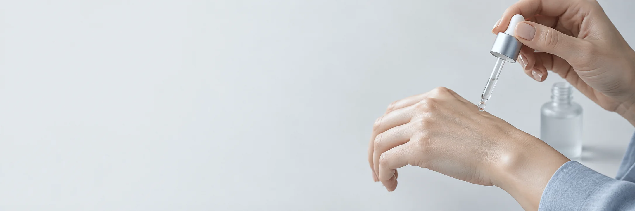 Hands applying a drop of clear serum from a glass dropper, beside a small bottle