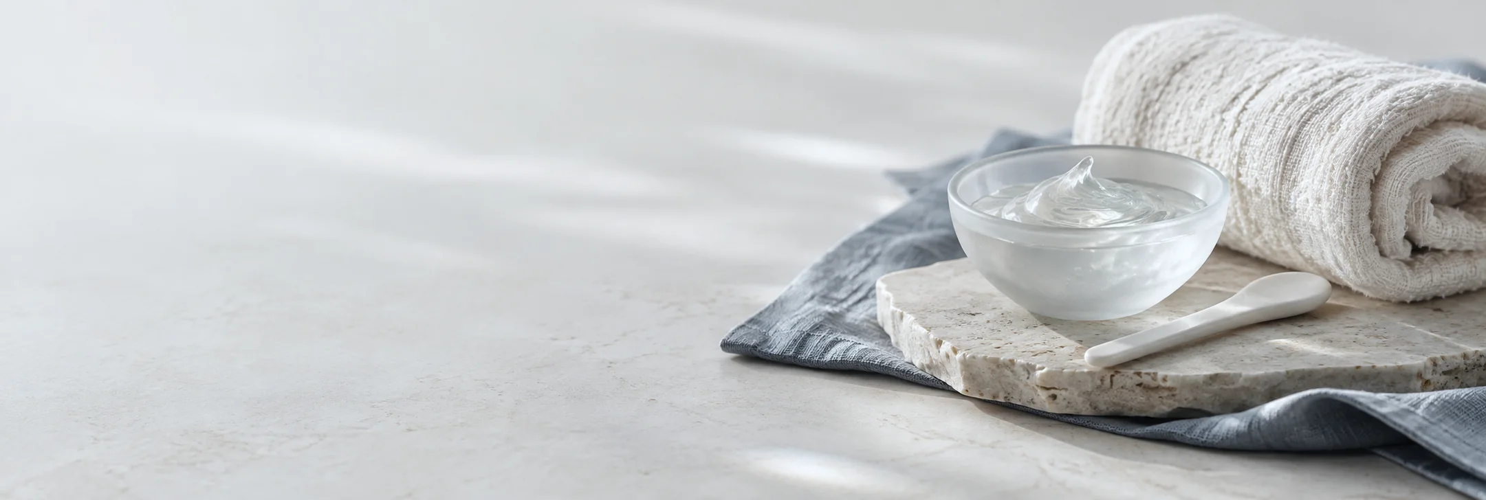A glass bowl of skincare gel with a spatula on a stone tray, beside a rolled white towel