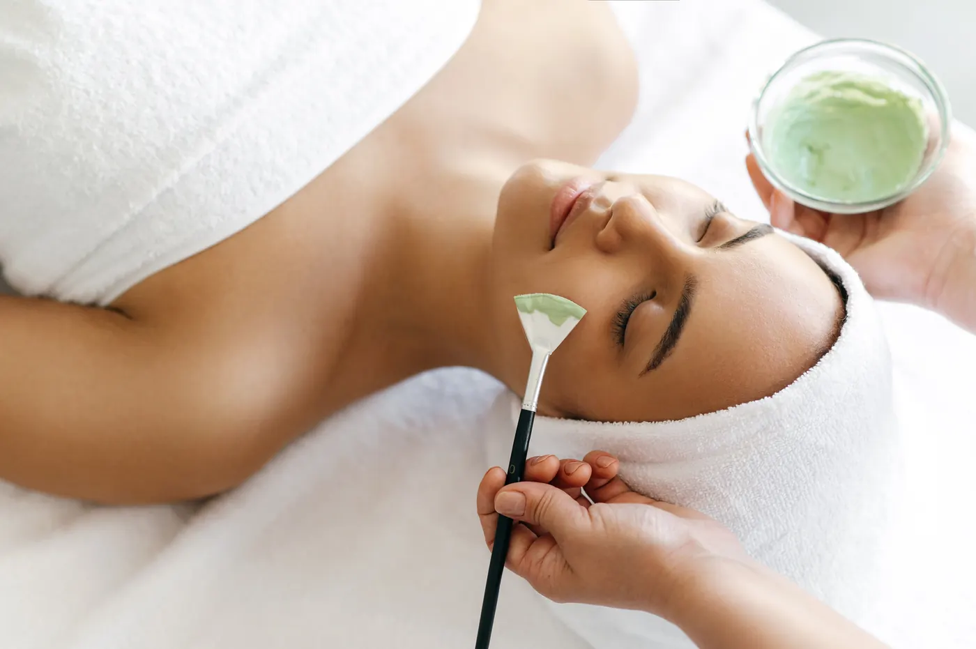 Facial treatment: a practitioner brushes a soft green mask onto a relaxed client’s cheek